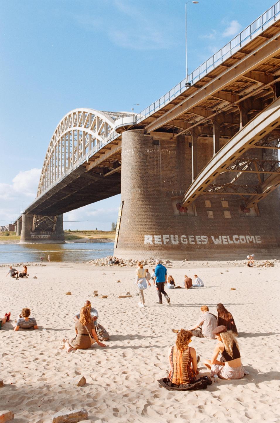 People relax on a sandy riverbank next to a large bridge with "REFUGEES WELCOME" painted on its base.