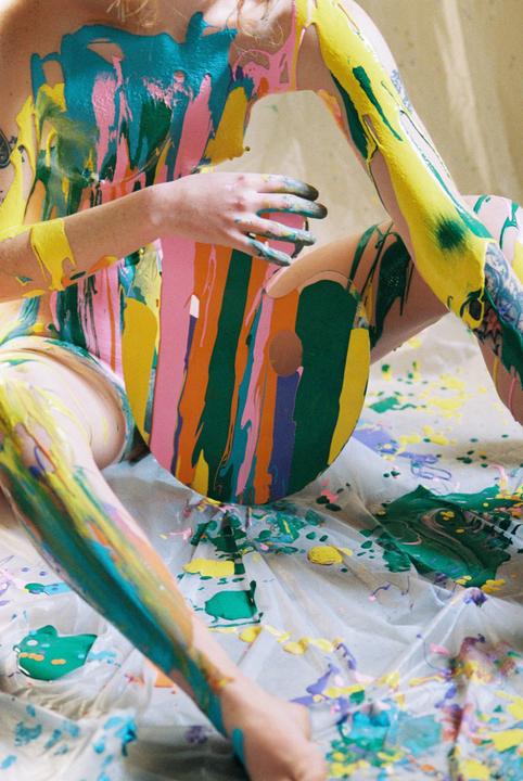 A person covered in colorful paint holds a paint palette while sitting on a paint-splattered sheet.