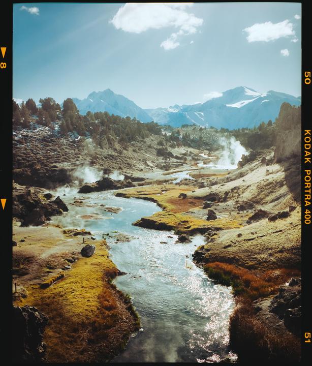 A winding river flows through a valley with steam rising from hot springs, surrounded by mountains and sparse vegetation.