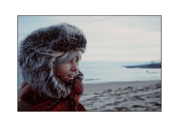 A woman wearing a fur hat and a red scarf looks out at the sea from a beach.