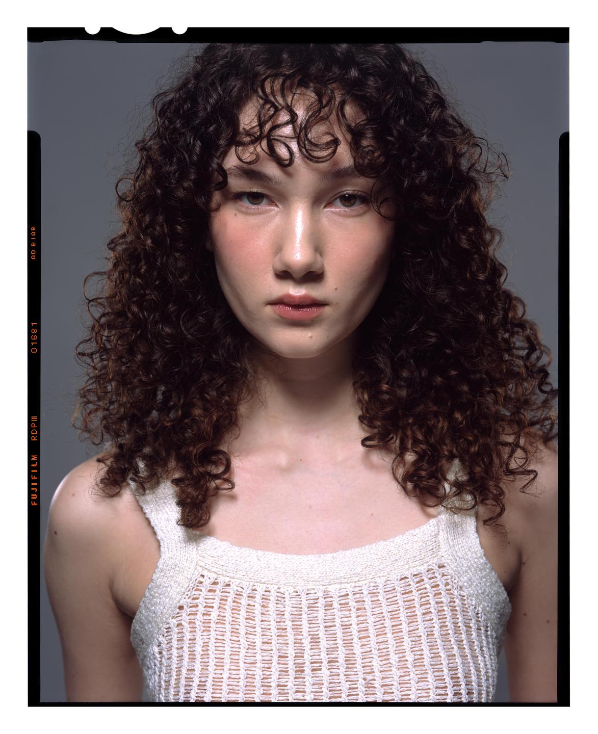 A woman with curly hair wears a white knit top and looks directly at the viewer.