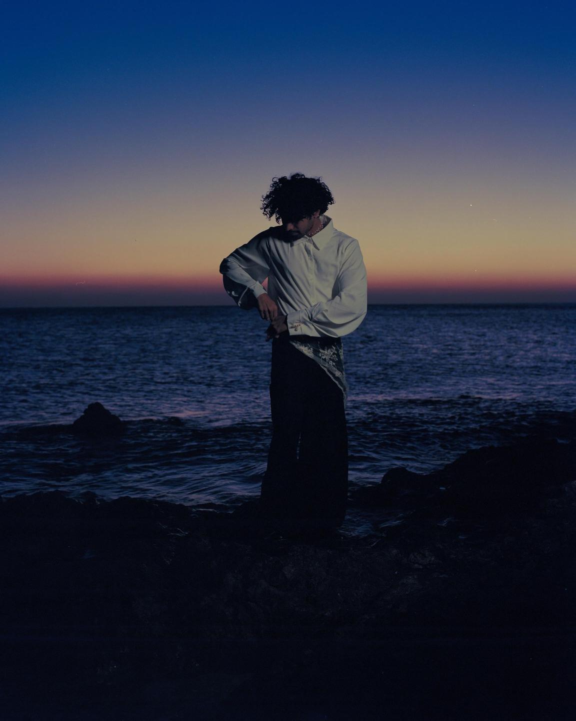 A man stands on a rocky shore with the ocean behind him at dawn.