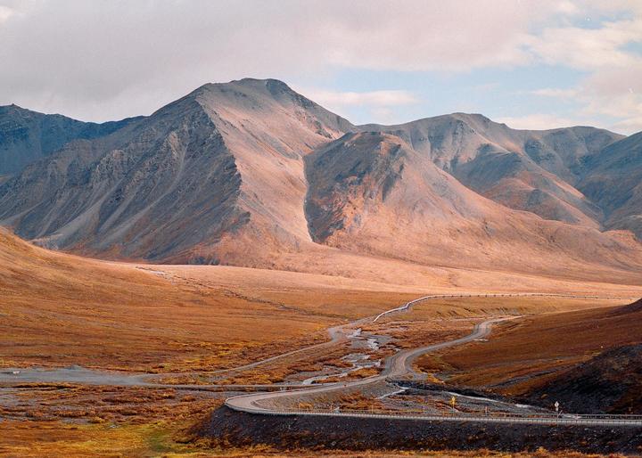 A winding road and pipeline cut through a vast, mountainous landscape with autumn foliage under a cloudy sky.