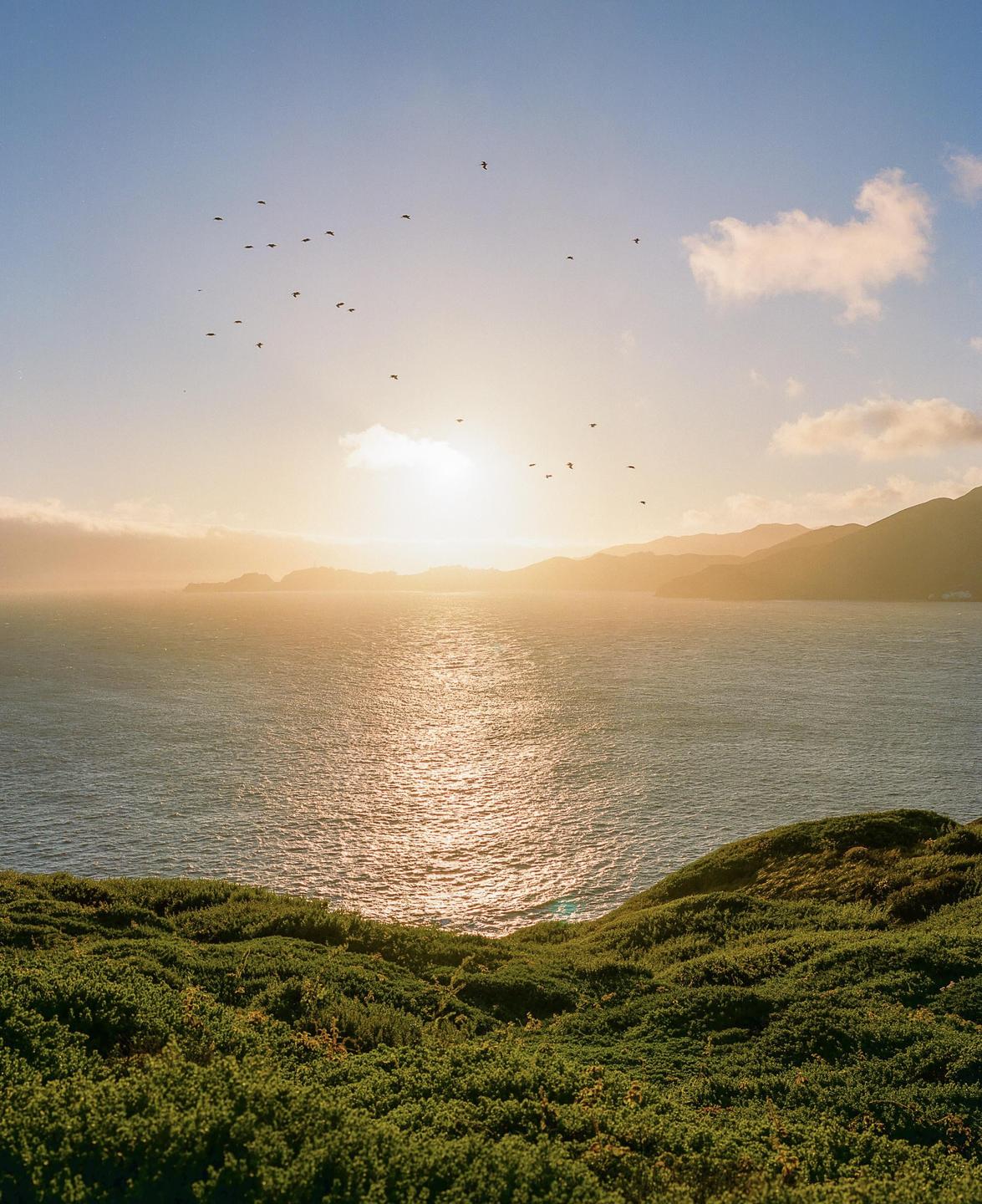 The sun sets over a body of water with hills in the distance and birds flying in the sky.