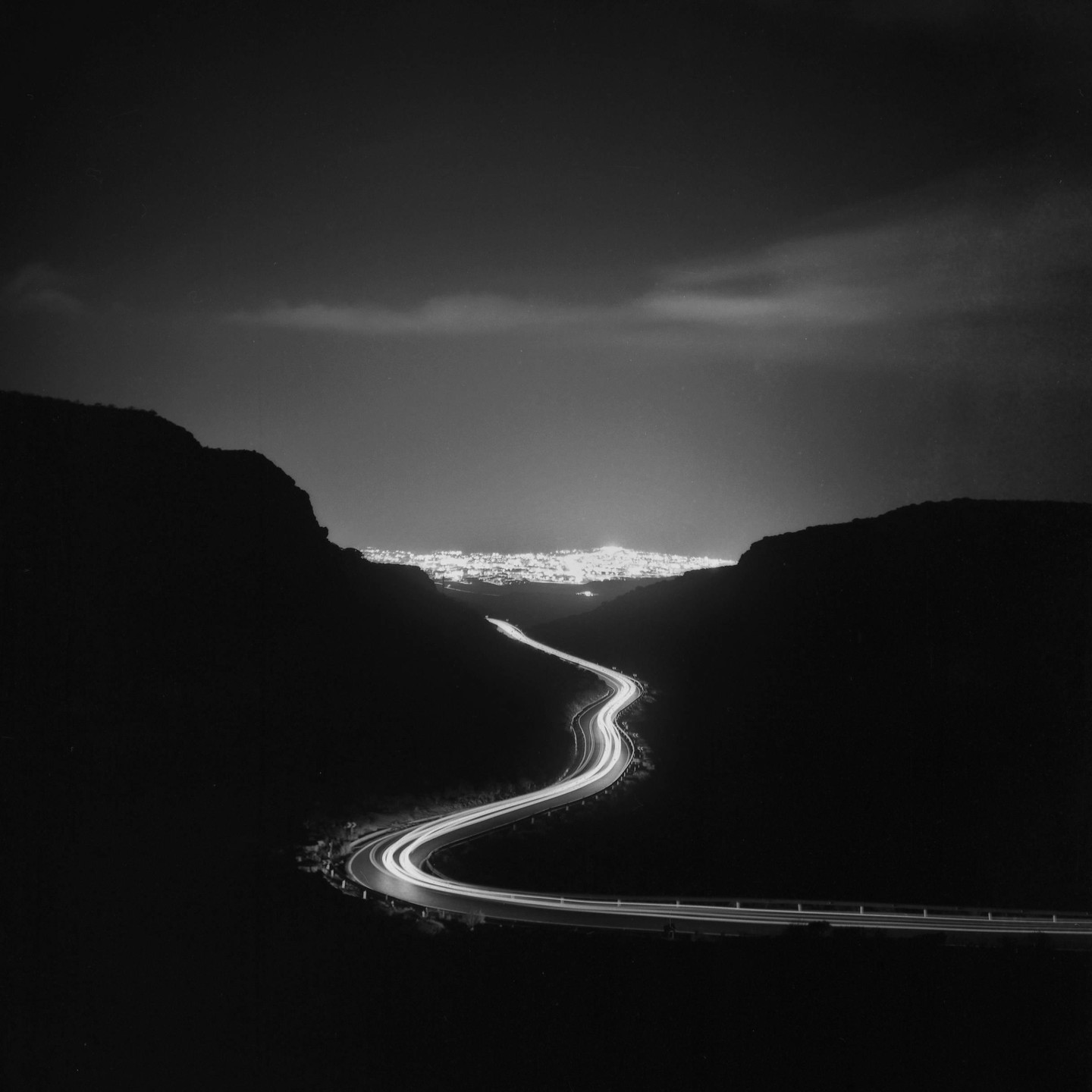 A winding road with light trails leads to a brightly lit city in the distance, framed by dark hills under a night sky.