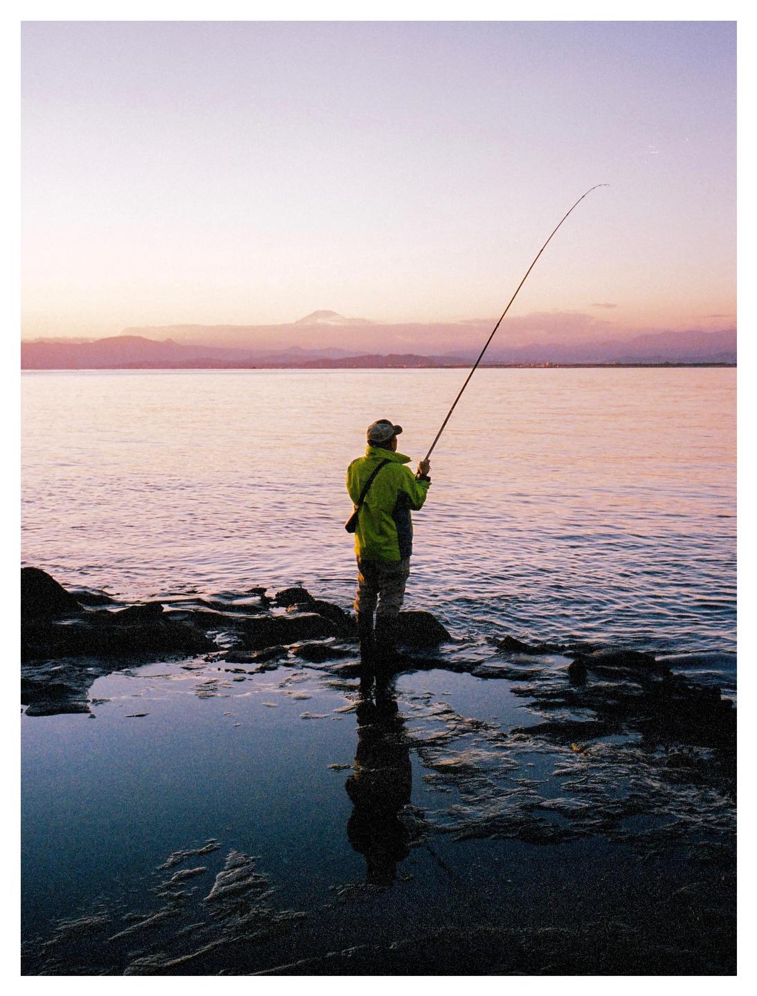 A man stands in shallow water on a rocky shore, fishing with a rod at sunset, with a mountain in the distance.
