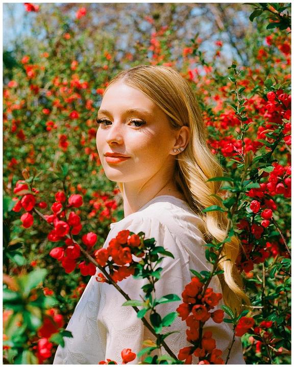 A woman with long blonde hair stands among red flowering bushes, looking over her shoulder at the viewer.