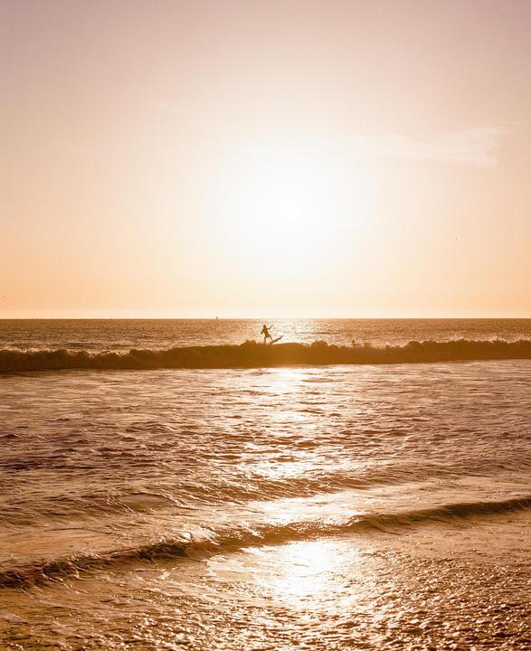 A surfer rides a wave at sunset, silhouetted against the bright sky.