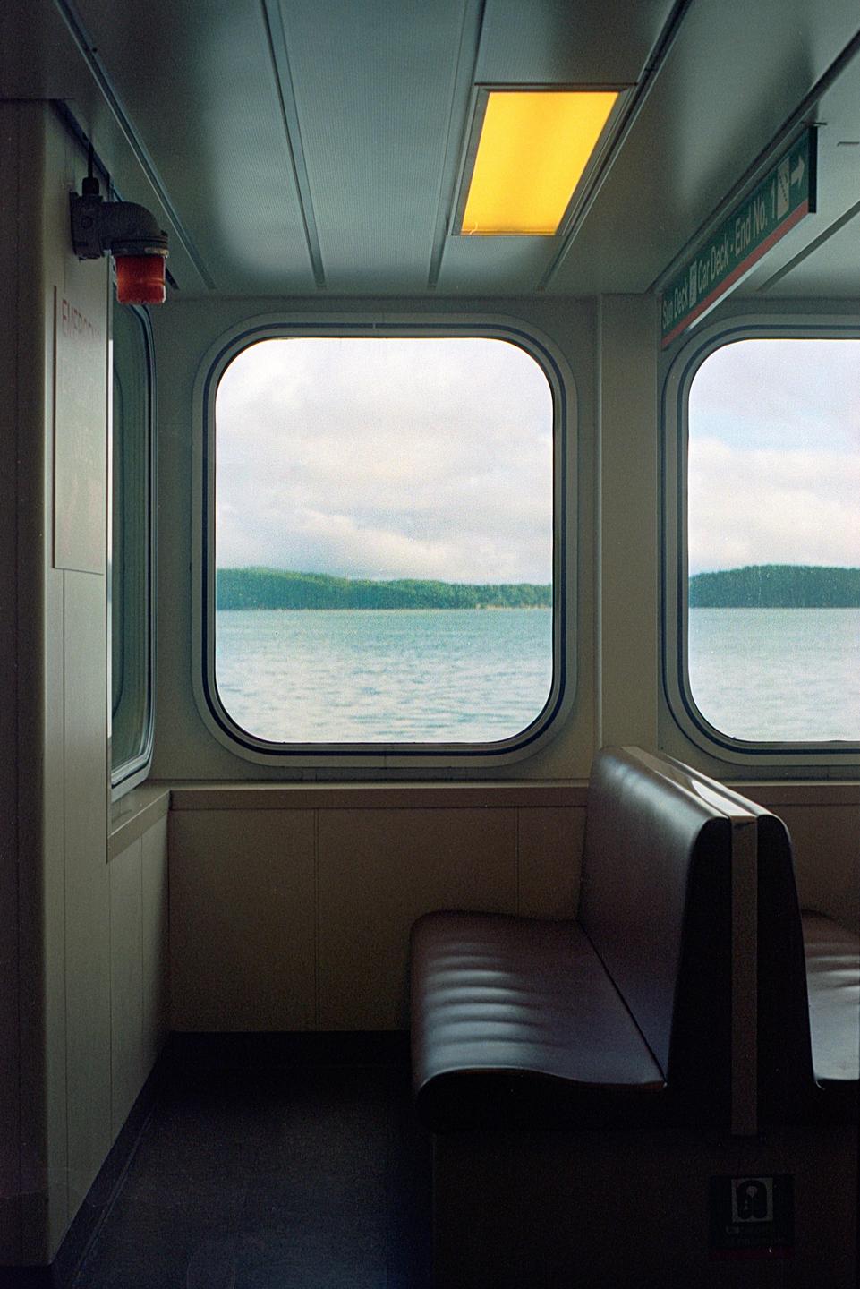 A ferry interior with a brown bench seat looks out two windows at a body of water and a distant forested shore under a cloudy sky.