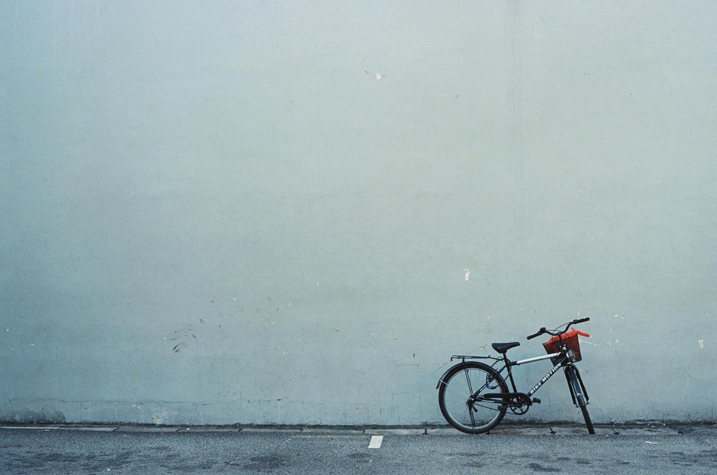 A bicycle with a red basket is parked against a light blue wall on an asphalt street.