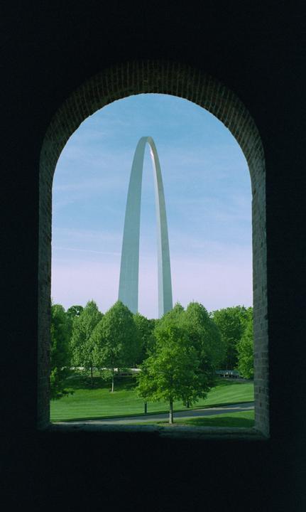 The Gateway Arch is visible through a dark brick archway, surrounded by green trees and grass under a clear blue sky.