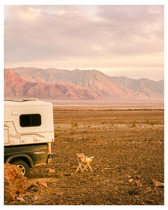 A truck camper is parked in a vast desert landscape with mountains in the distance under a cloudy sky.