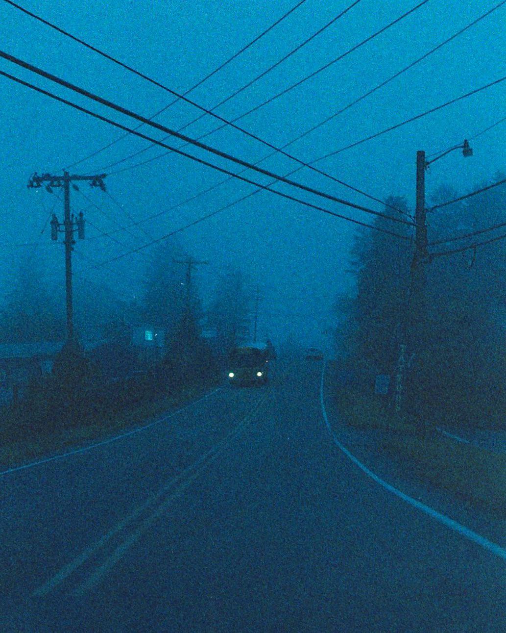 A car with its headlights on drives down a foggy road lined with utility poles and trees.