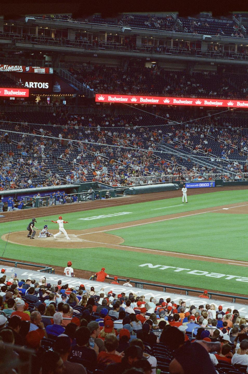 A baseball game is underway in a stadium filled with spectators, with players on the field and advertisements visible on the upper decks.