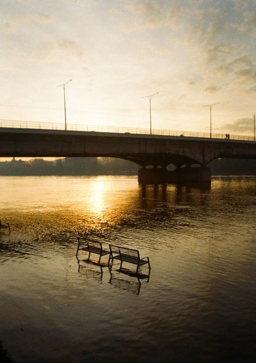 Two submerged benches sit in a body of water with a bridge and golden hour sky in the background.