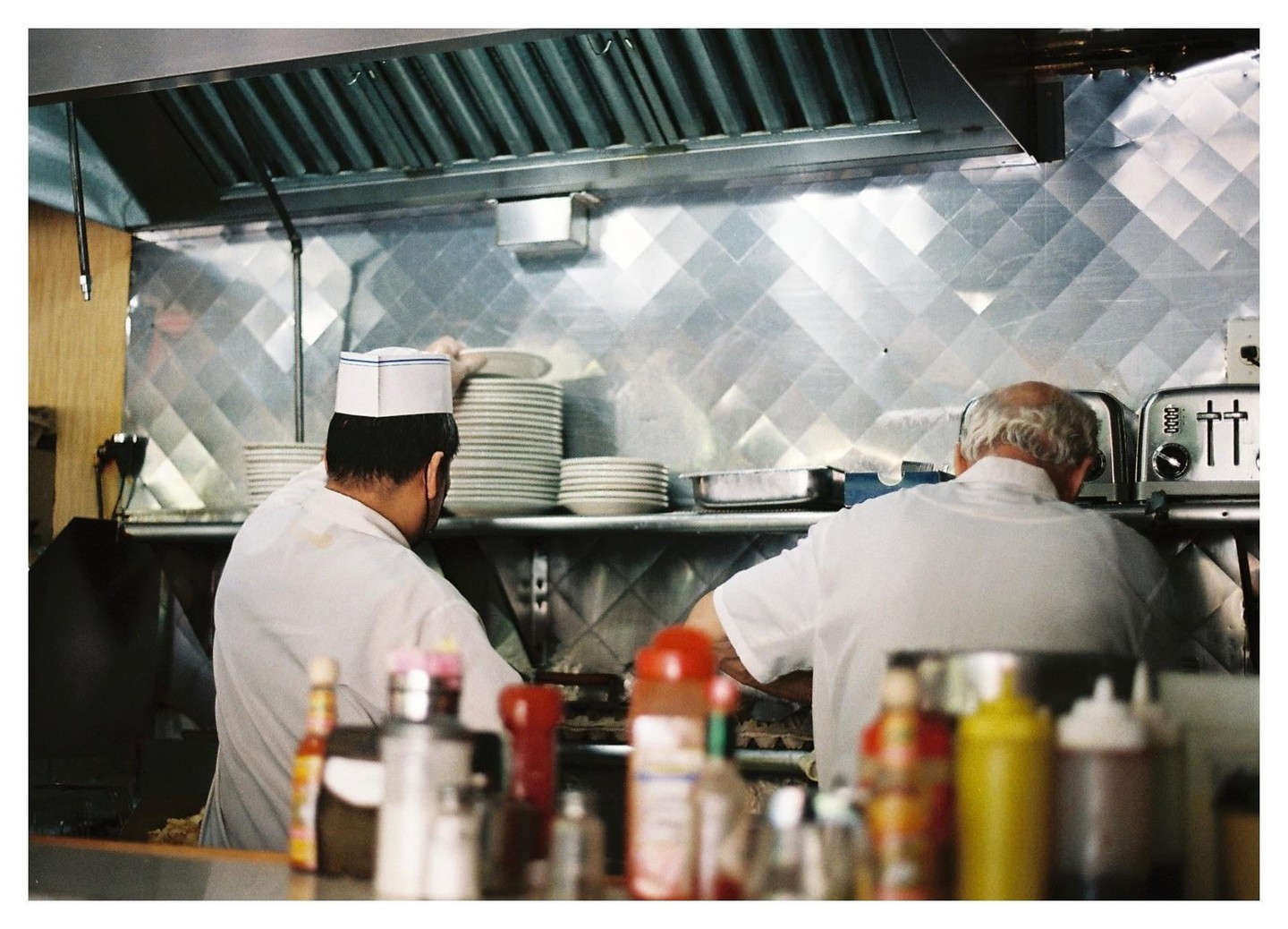 Two men work in a diner kitchen with stainless steel walls and a shelf of stacked plates.