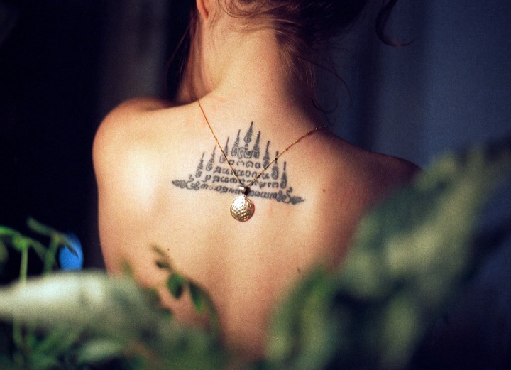 A woman's back is visible, adorned with a tattoo and a necklace with a round pendant, with green foliage in the foreground.