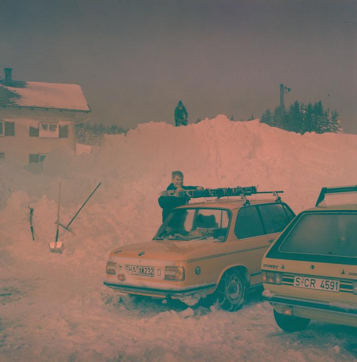 A man stands next to a yellow car with skis on its roof, surrounded by deep snow and another person in the distance.