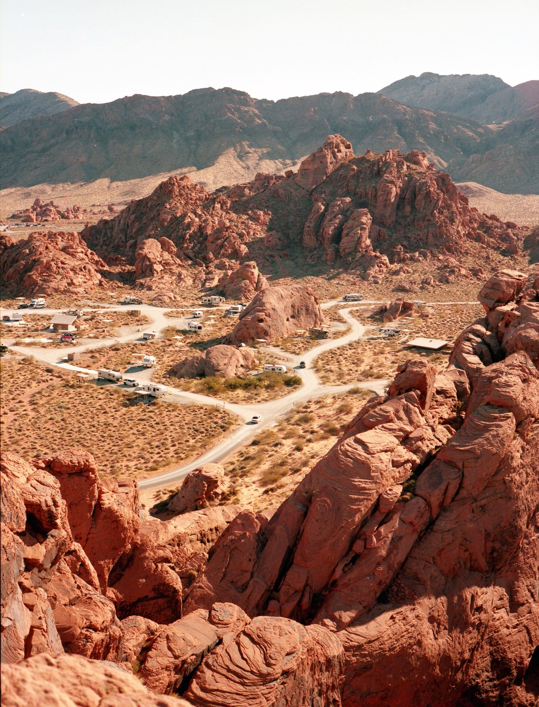 A high-angle landscape shows a desert campground nestled among red rock formations with mountains in the distance.