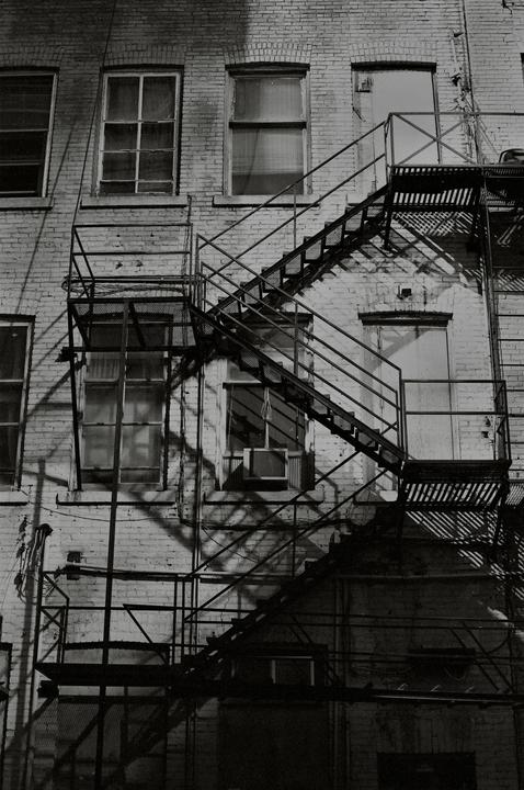 A fire escape with diagonal shadows is attached to a brick building with multiple windows.