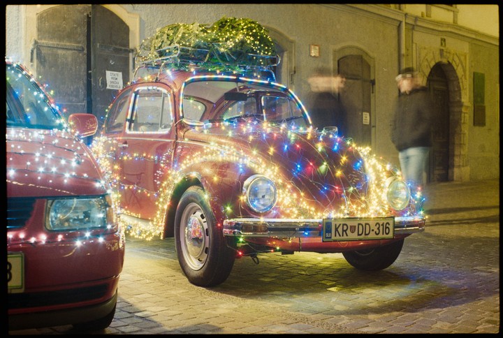A red volkswagen beetle covered in colorful christmas lights is parked on a cobblestone street at night with a christmas tree on its roof.