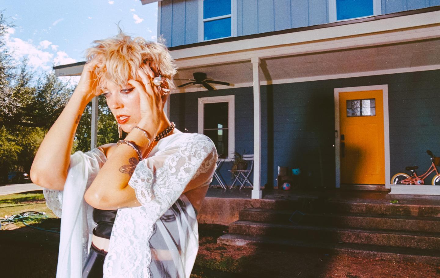 A woman with short blonde hair and a white lace top stands in front of a blue house with a yellow door.