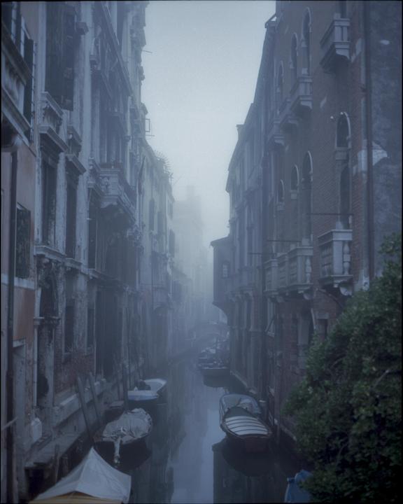 A narrow canal with boats is visible between tall buildings on a hazy day in Venice.