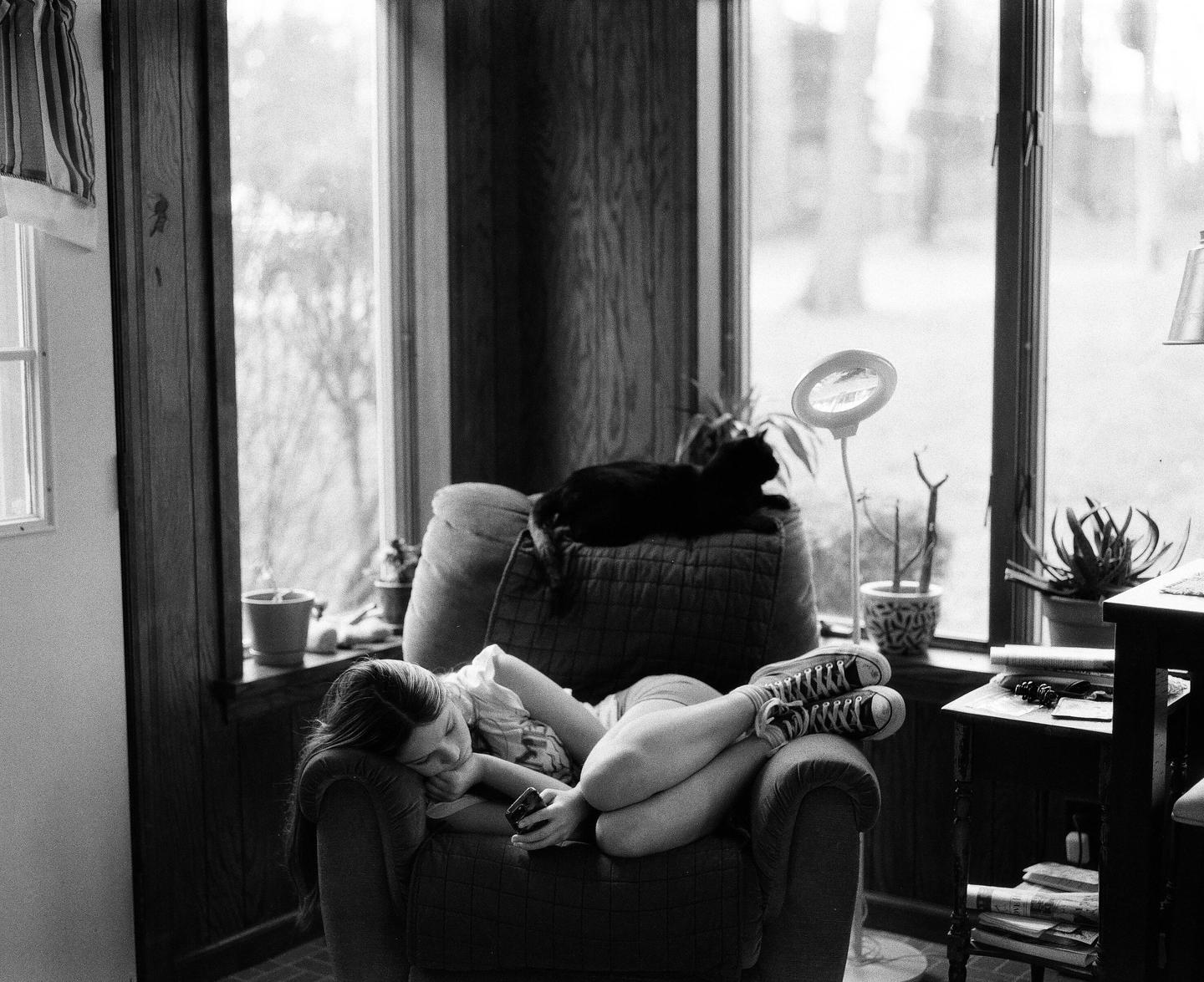 A woman reclines in an armchair with a cat on the back of the chair next to a window.