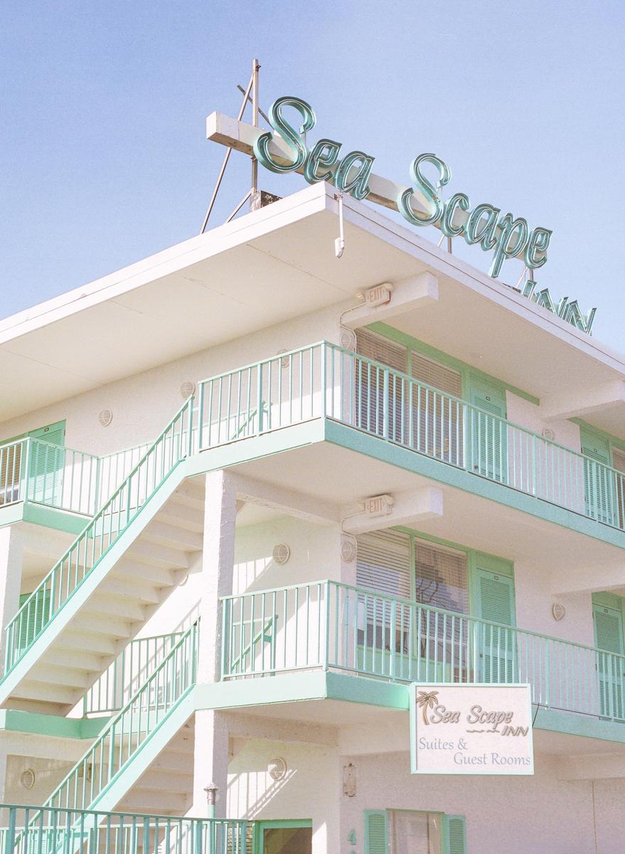 A white building with mint green railings and stairs has a "Sea Scape Inn" sign on the roof and another sign for "Sea Scape Suites & Guest Rooms" on the lower level.