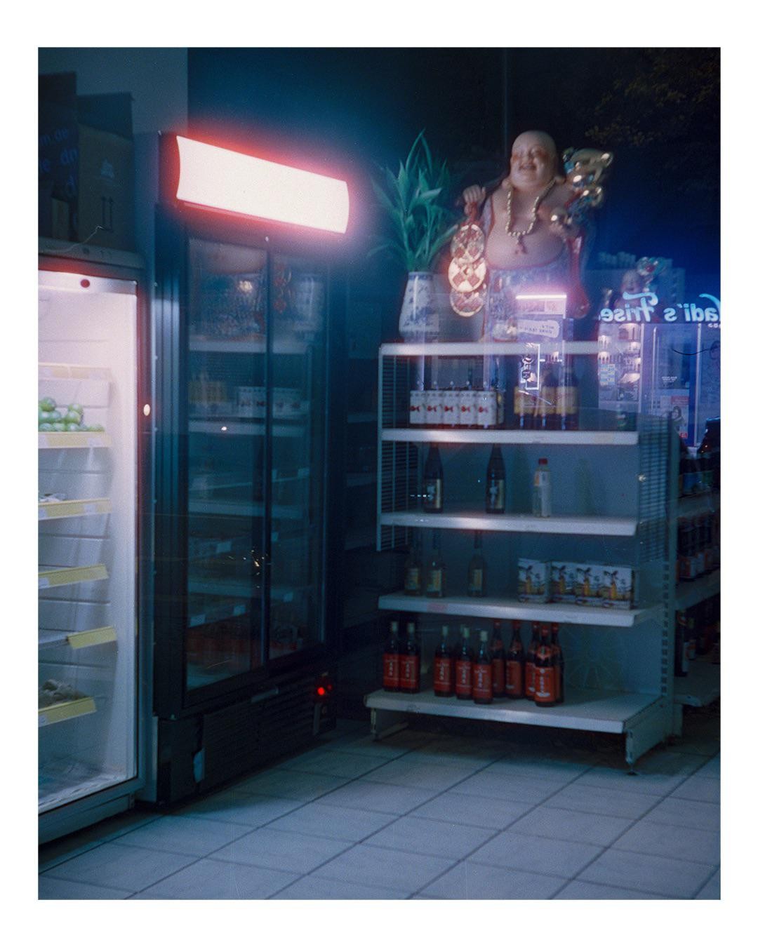 A Buddha statue stands on a shelf next to a neon sign and a refrigerator in a store at night.