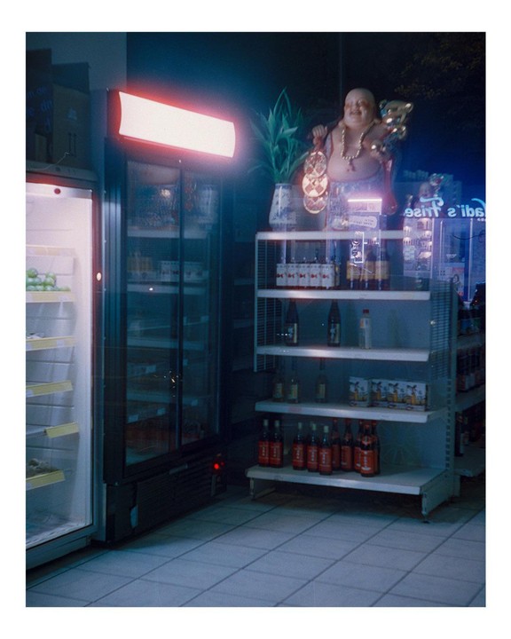 A Buddha statue stands on a shelf next to a neon sign and a refrigerator in a store at night.