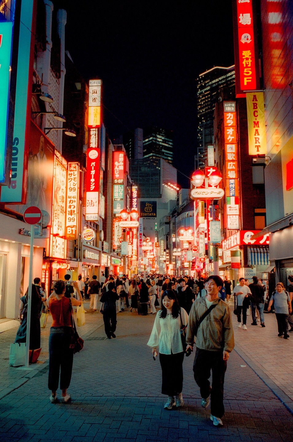 A couple walks down a busy street filled with neon signs and people at night.