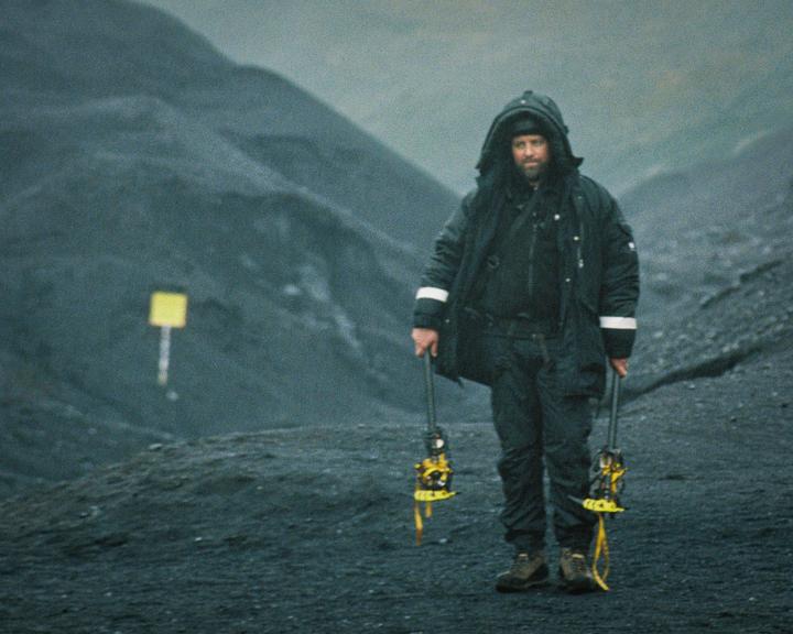 A man in a hooded jacket and boots stands on a dark, rocky landscape holding two ice axes.