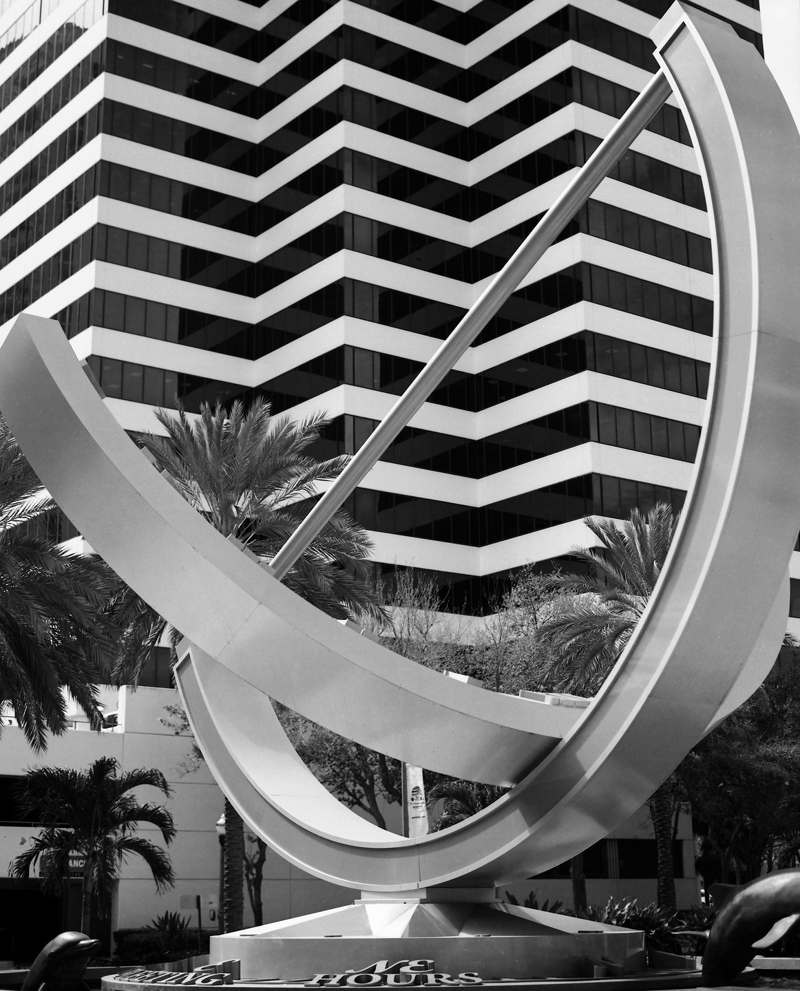 A large, modern sundial sculpture stands in front of a building with a zig-zag pattern of windows and white panels, with palm trees visible around the base.