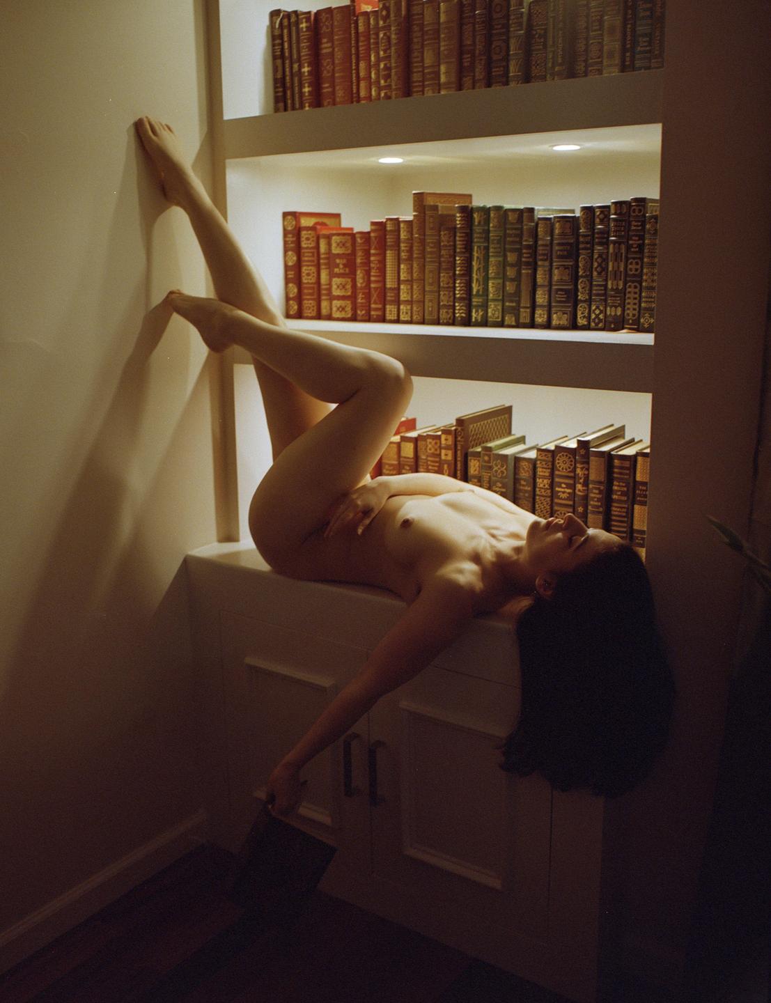 A nude woman reclines on a cabinet with her legs up against a wall, surrounded by bookshelves.