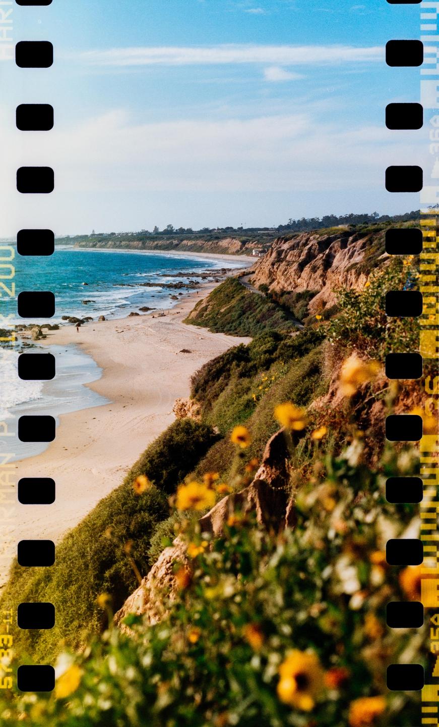 A sandy beach curves along a coastline with cliffs and green vegetation under a blue sky.