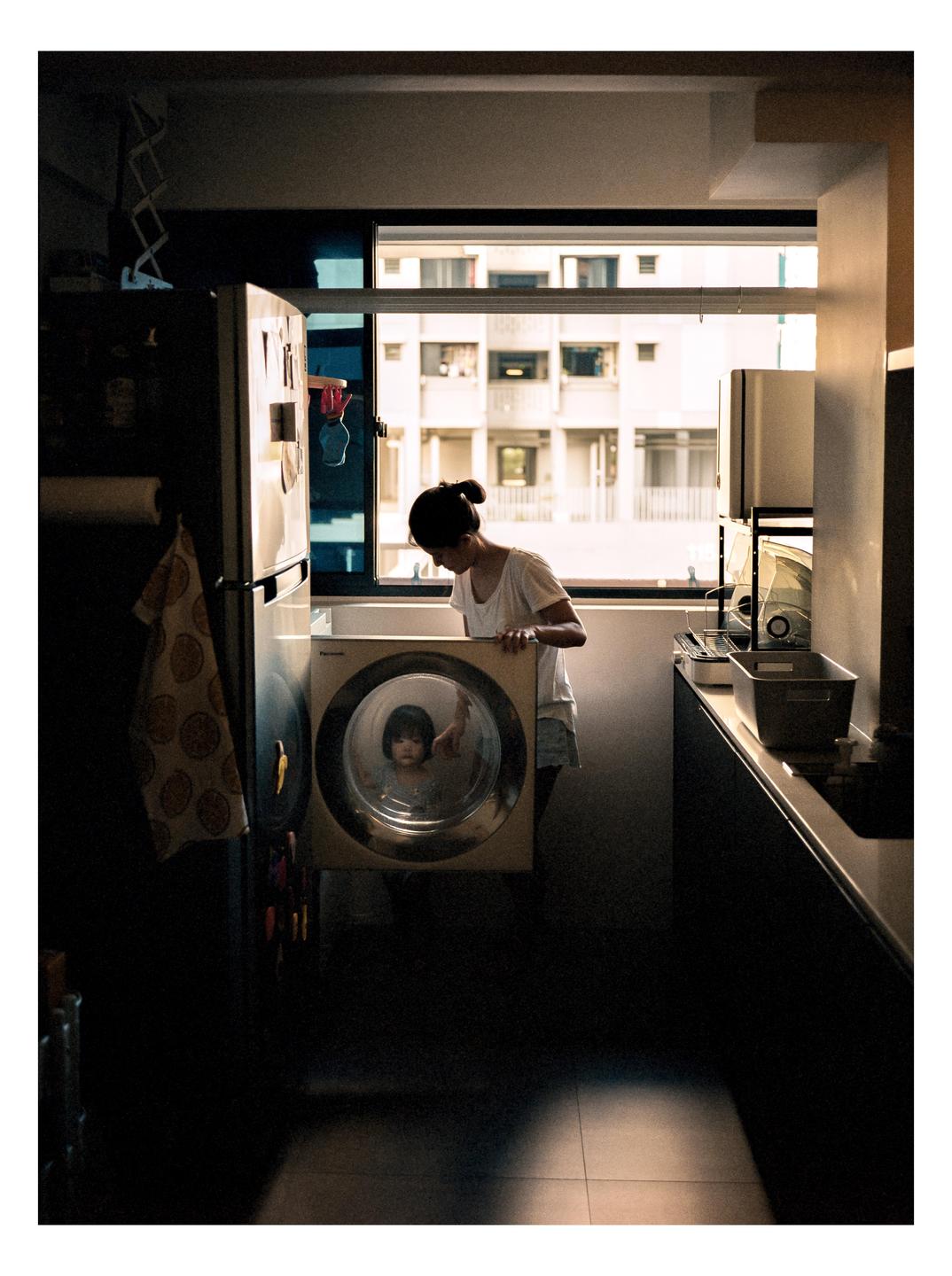 A woman looks into a washing machine where a child is visible through the door, in a kitchen with a window looking out onto apartment buildings.
