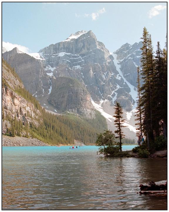 A turquoise lake is surrounded by a forest and snow-capped mountains under a clear sky.
