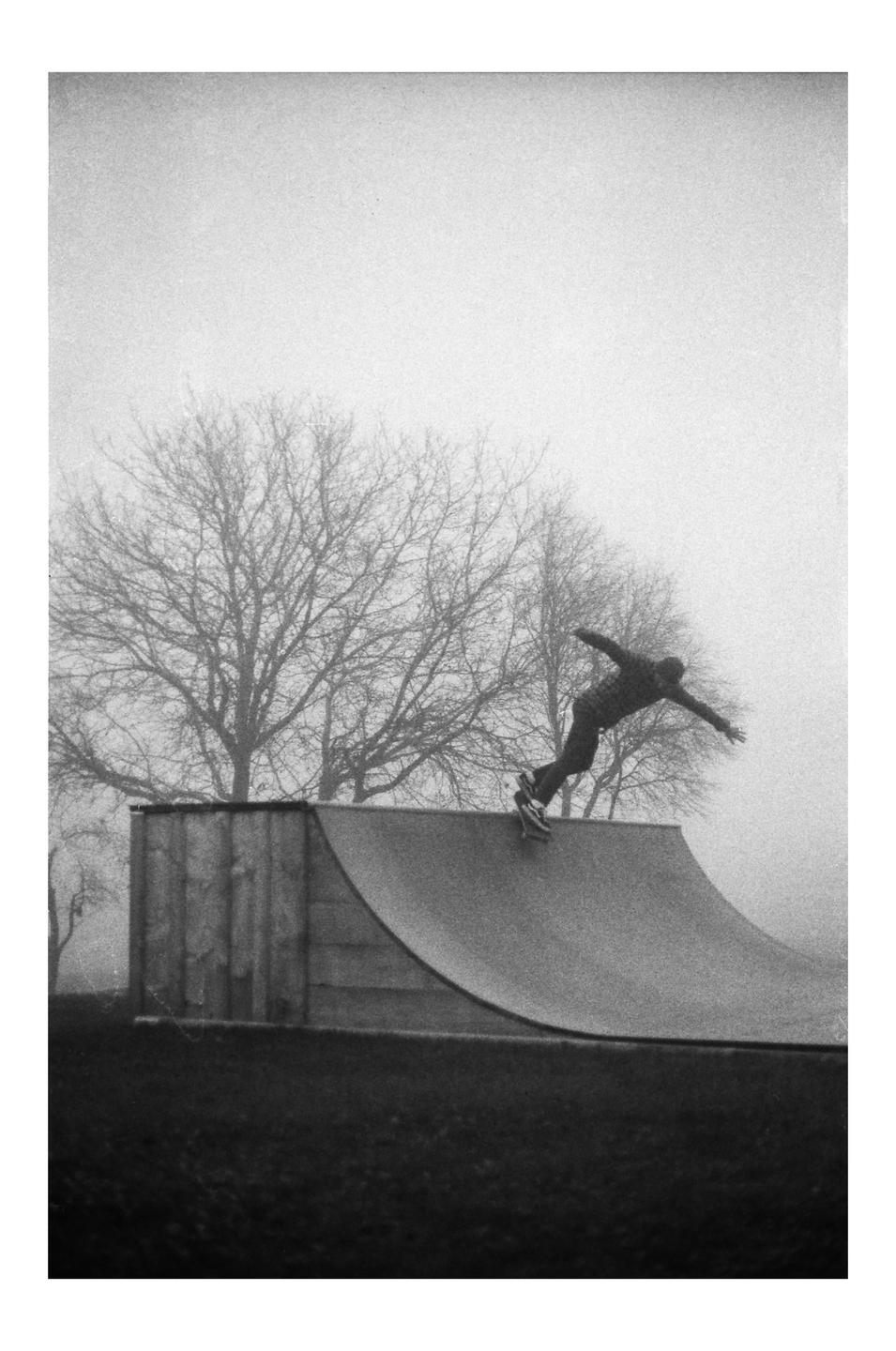A person on a skateboard is captured mid-air on a ramp, with bare trees and a hazy sky in the background.
