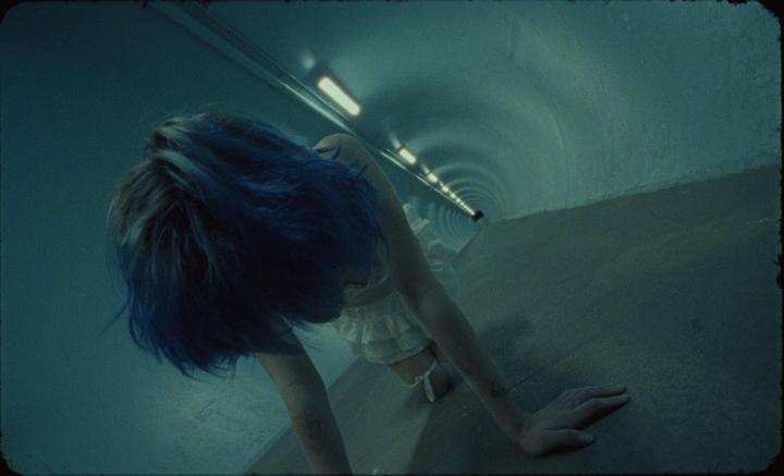 A woman with blue hair crawls on a concrete surface in a tunnel.