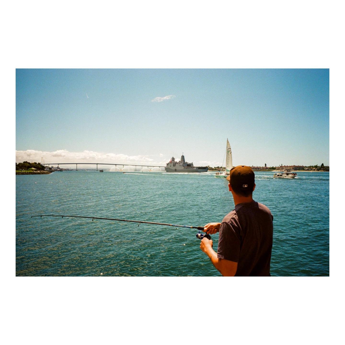 A man fishes from a pier with a large ship, sailboat, and bridge in the background.