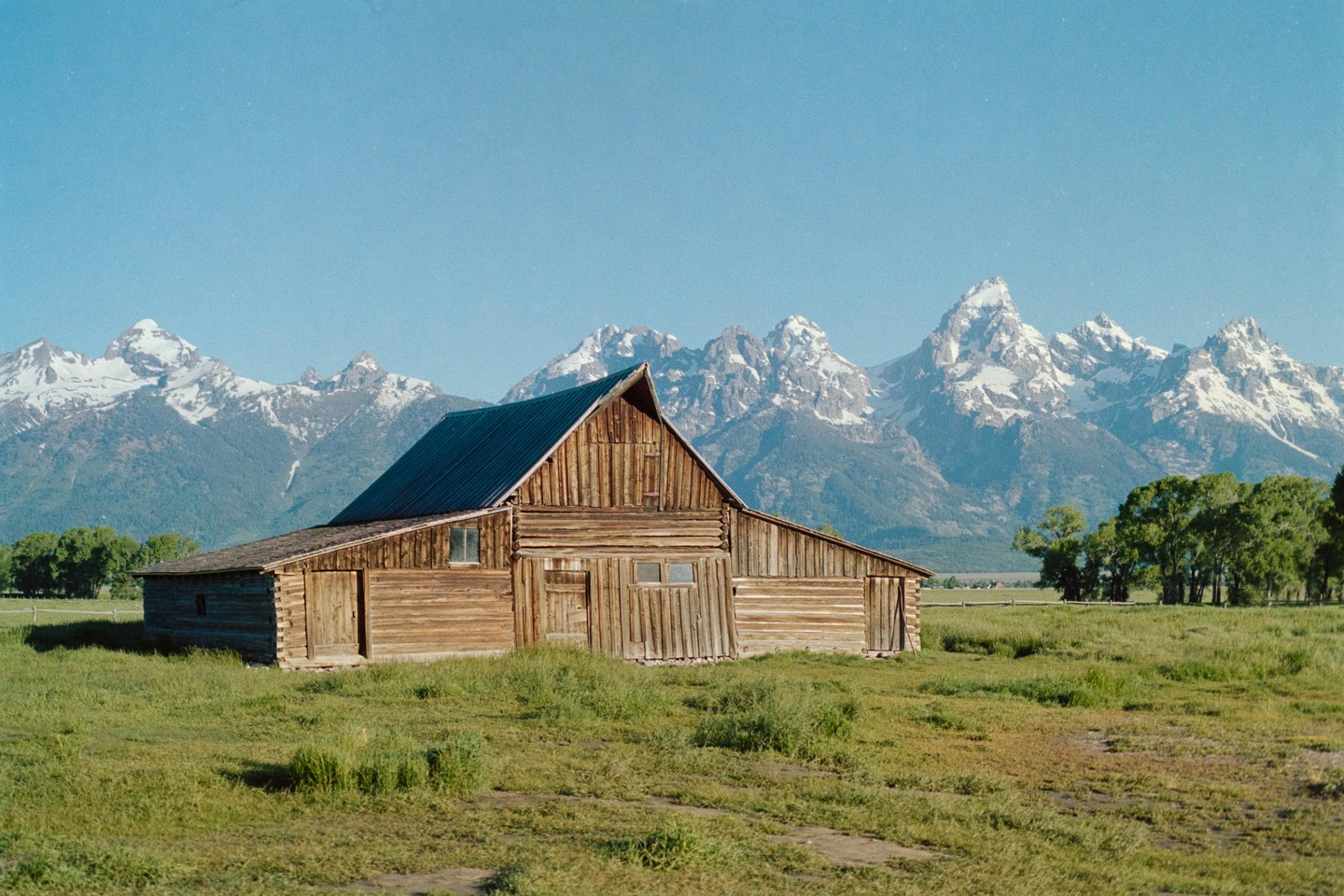 A wooden barn stands in a grassy field with snow-capped mountains in the background under a clear sky.