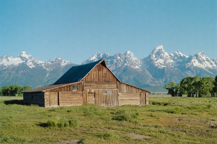 A wooden barn stands in a grassy field with snow-capped mountains in the background under a clear sky.