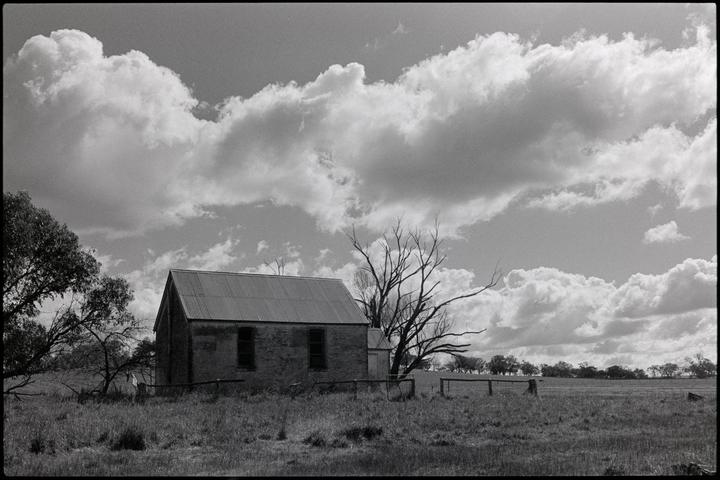 An old brick building with a corrugated iron roof stands in a grassy field under a cloudy sky.