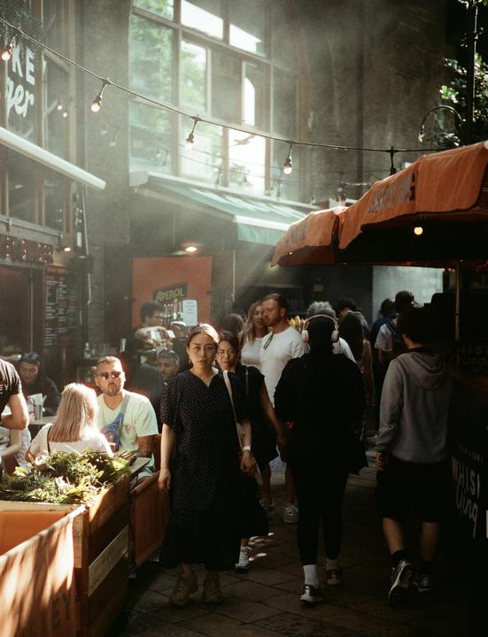 A woman in a dark dress walks through a crowded market with string lights overhead and sunlight streaming through a window.