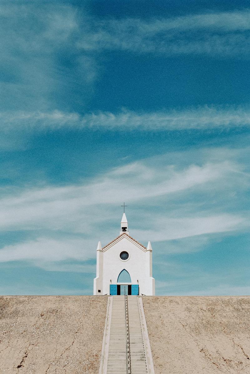 A white church with blue doors sits atop a hill, with a long staircase leading up to it under a blue sky with wispy clouds.