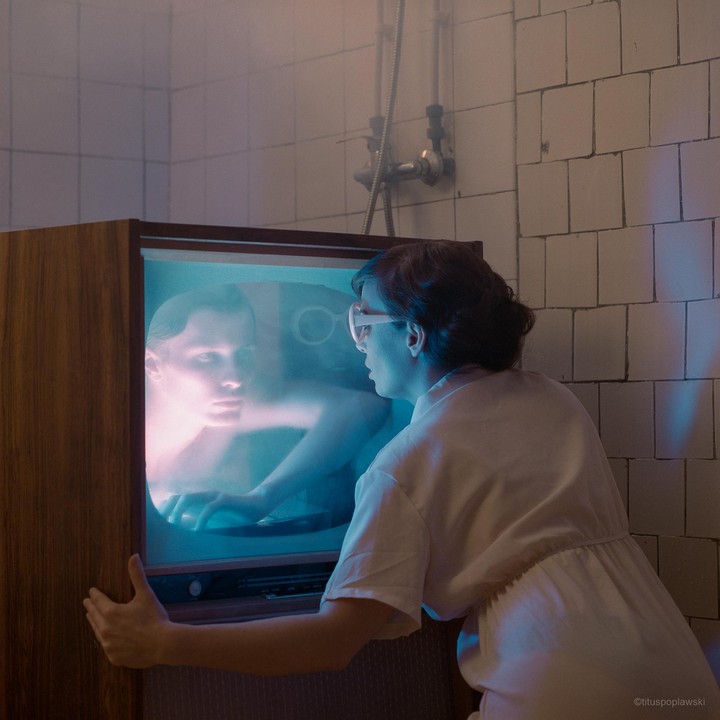 A woman in a white dress and glasses looks into a television screen showing a man's face.