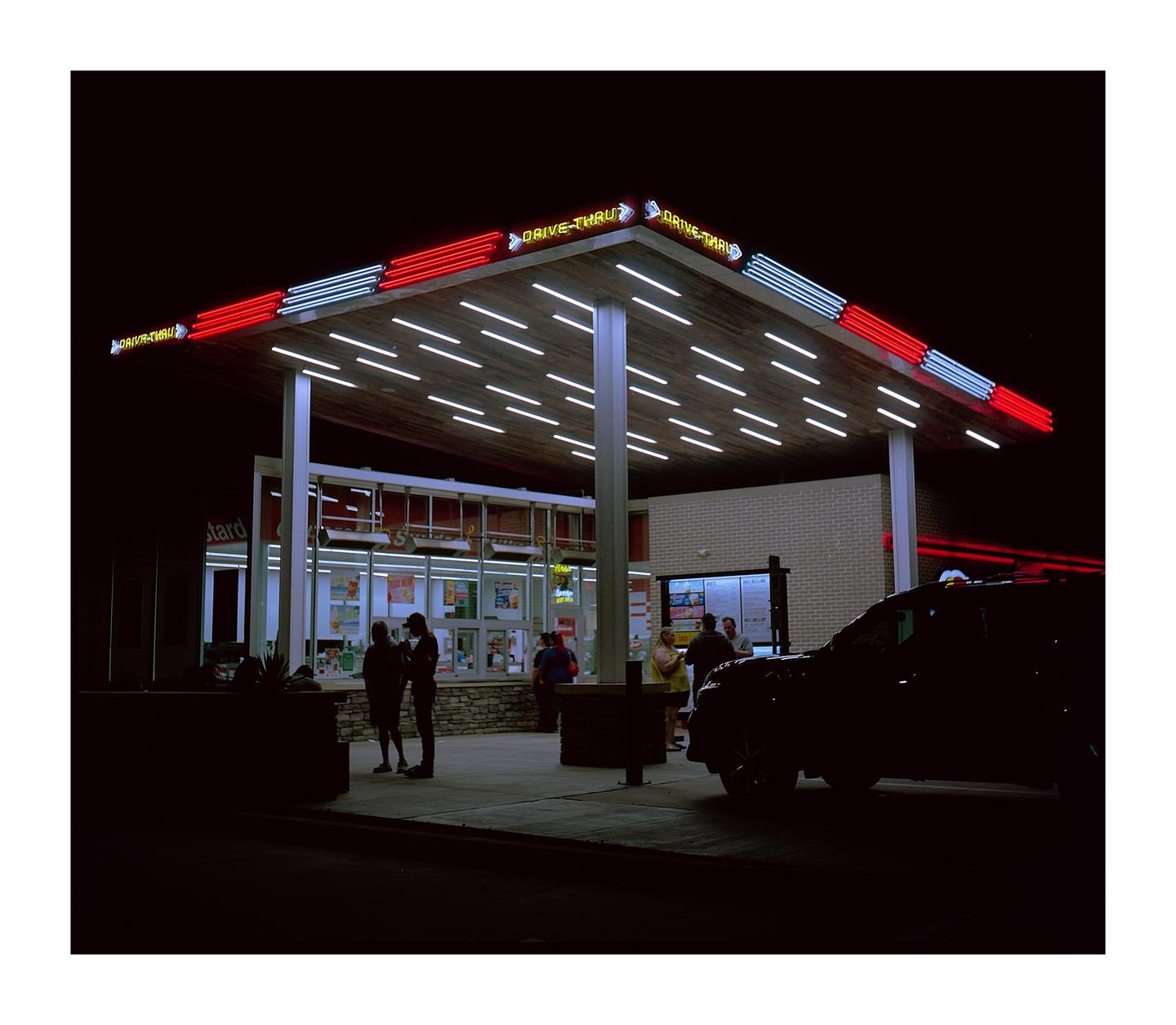 A drive-thru restaurant with neon lights is illuminated at night, with people and a car visible.