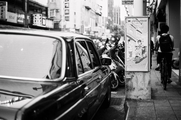 A black car is parked on a city street next to a row of scooters and a person on a bicycle.