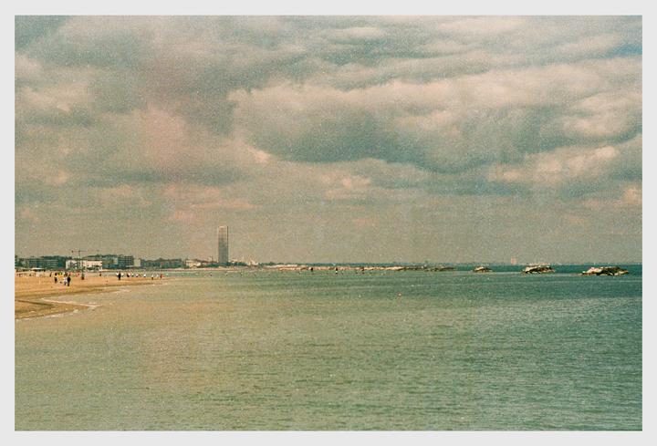 A beach with a city and a tall building in the distance is visible under a cloudy sky.